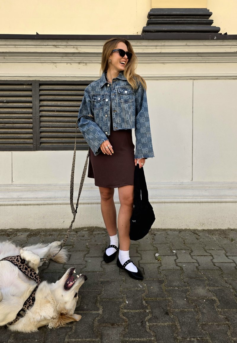 Veste en denim avec motif de logo, robe marron, chaussettes blanches, chaussures noires avec accents. Un chien est allongé à proximité sur des pavés.