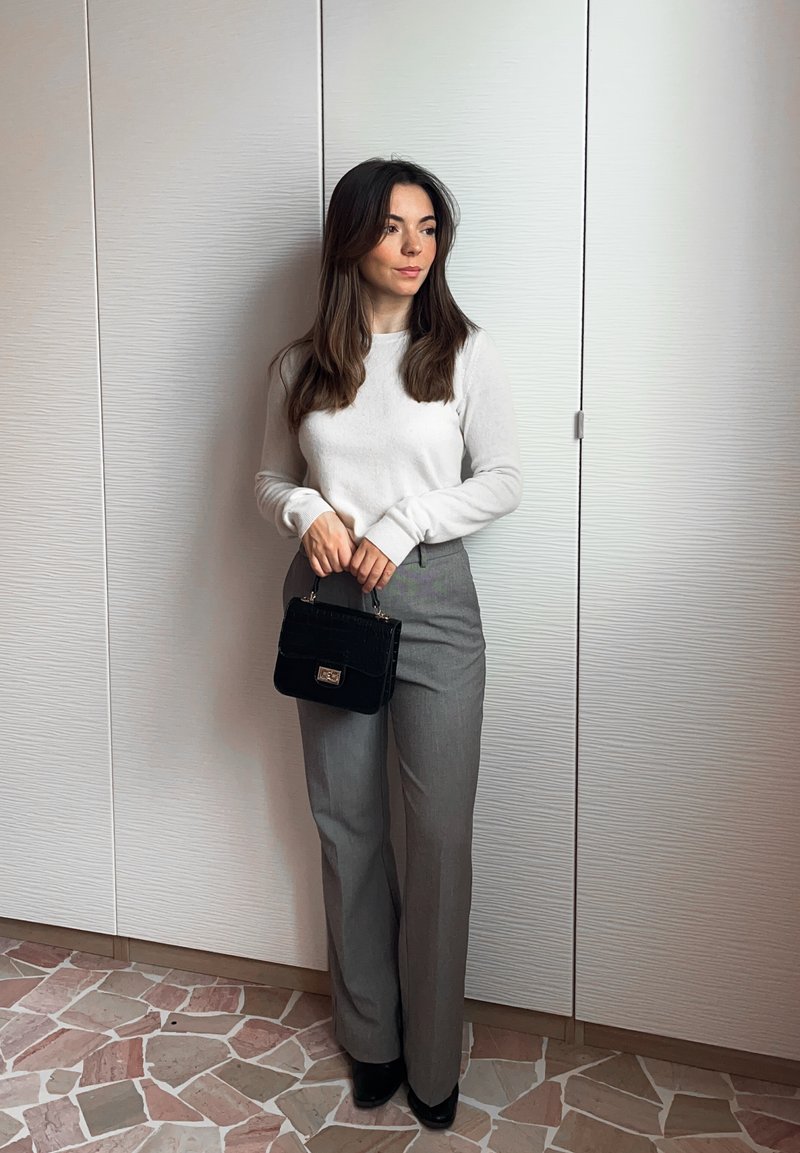 Pantaloni gri deschis asortați cu un top alb cu mânecă lungă, ținând o geantă mică neagră, stând pe un podea din piatră cu model.