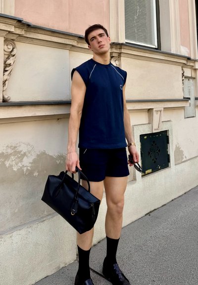 Camiseta sin mangas de color navy con acentos en gris claro, combinada con pantalones cortos oscuros. Sosteniendo un bolso de cuero negro, lleva calcetas altas negras y zapatos.
