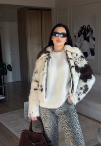 Giacca di pelliccia sintetica con motivi maculati marroni e bianchi, abbinata a un maglione chiaro e pantaloni con stampa animale, mentre tiene una borsa marrone.