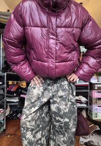 Osoba oblečená v lesklej fialovej pufre a abstraktných šedých maskáčových nohaviciach stojí v preplnenej miestnosti so poličkami a oblečením.