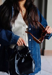 Black, soft leather handbag with a ruched handle, paired with a denim jacket and a white top. Holding brown tortoiseshell sunglasses.