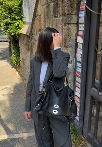 Fekete bőr kézitáska ezüstszínű kiegészítőkkel, laza kialakítással és szegecselt díszítéssel. Szürke kockás blézer és fehér felső, amelyet egy személy visel.