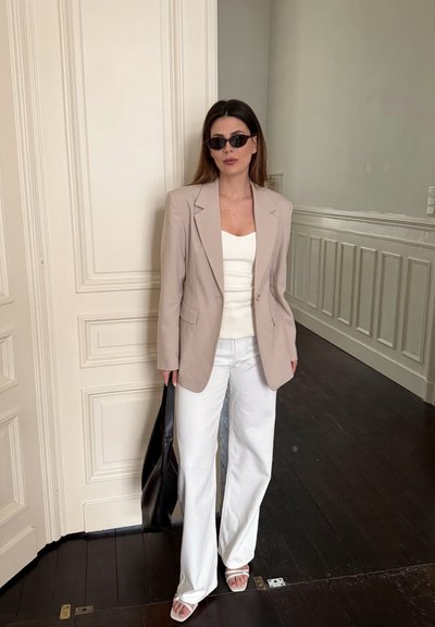 Mujer con blazer beige, top blanco, pantalones blancos, gafas de sol y sandalias blancas, sosteniendo un bolso negro, de pie en el interior cerca de una puerta blanca.