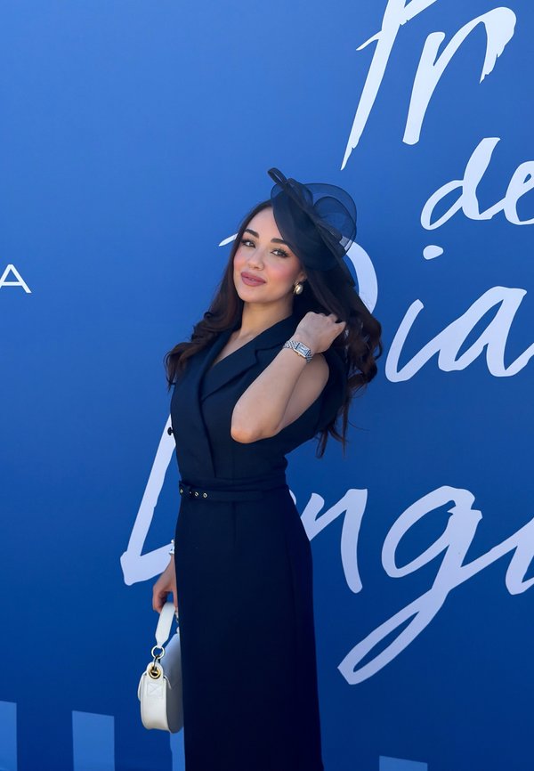 Femme en robe noire sans manches et chapeau posant avec un sac à main blanc sur un fond bleu avec du texte blanc en cursive.