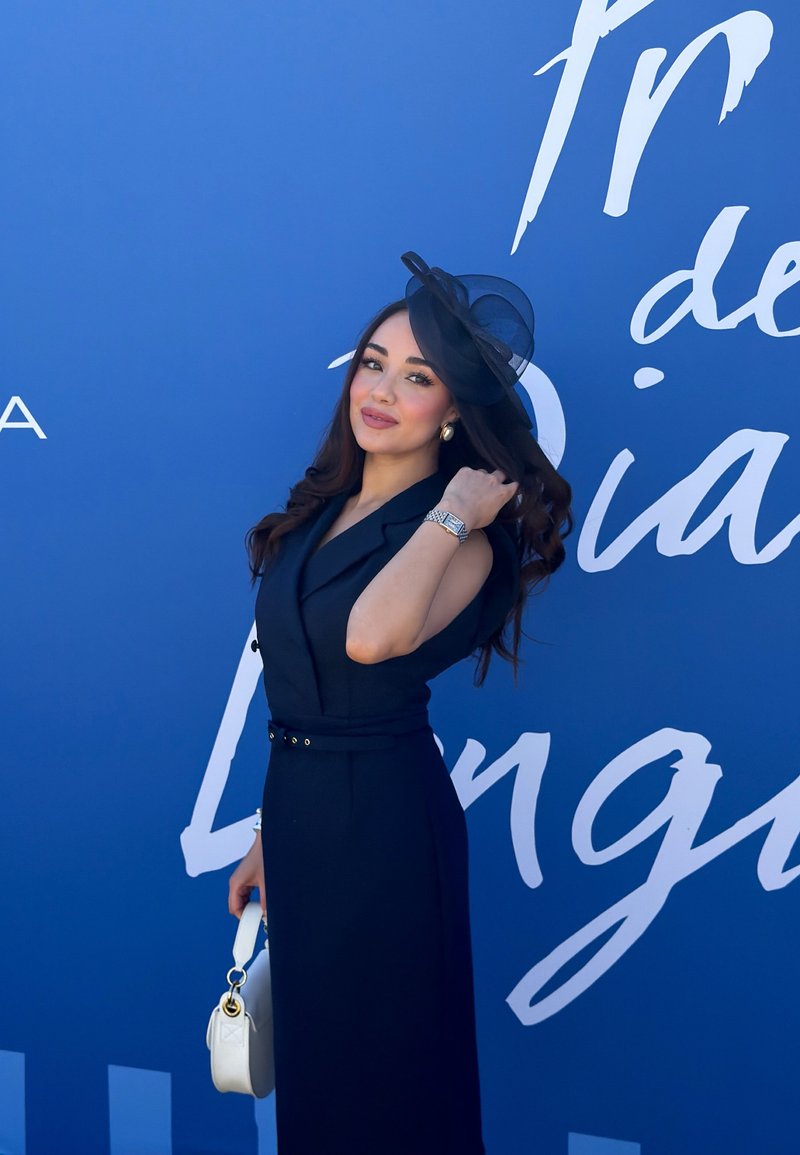 Femme en robe noire sans manches et chapeau posant avec un sac à main blanc sur fond bleu avec un texte blanc en cursive.