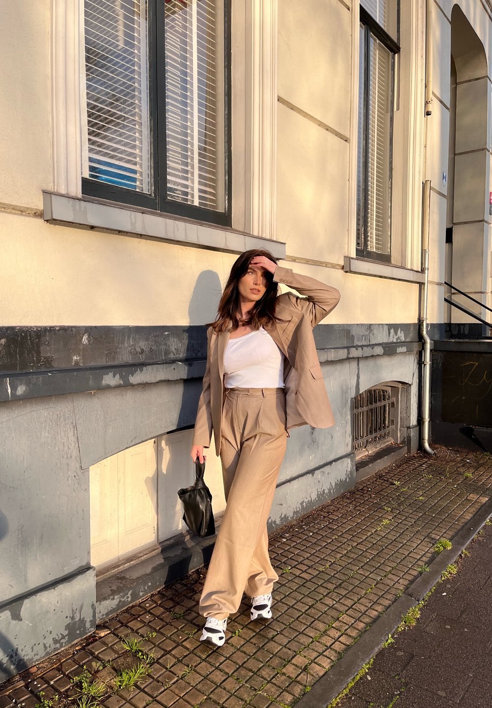 Costume beige en deux pièces avec pantalons à jambes larges et blazer ajusté, associé à un débardeur blanc et un sac à main noir. Chaussures de sport décontractées.