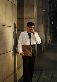 Λευκό κοντό σακάκι πάνω από μαύρη μπλούζα, μαύρο παντελόνι και καφέ δερμάτινη τσάντα, μπροστά σε μια υφασμάτινη τοιχογραφία από φυσική πέτρα με απαλό βραδινό φωτισμό.