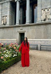 Rode linnenoutfit met een losse top en wijde broekspijpen. De achtergrond bevat stenen architectuur en kleurrijke bloemen.