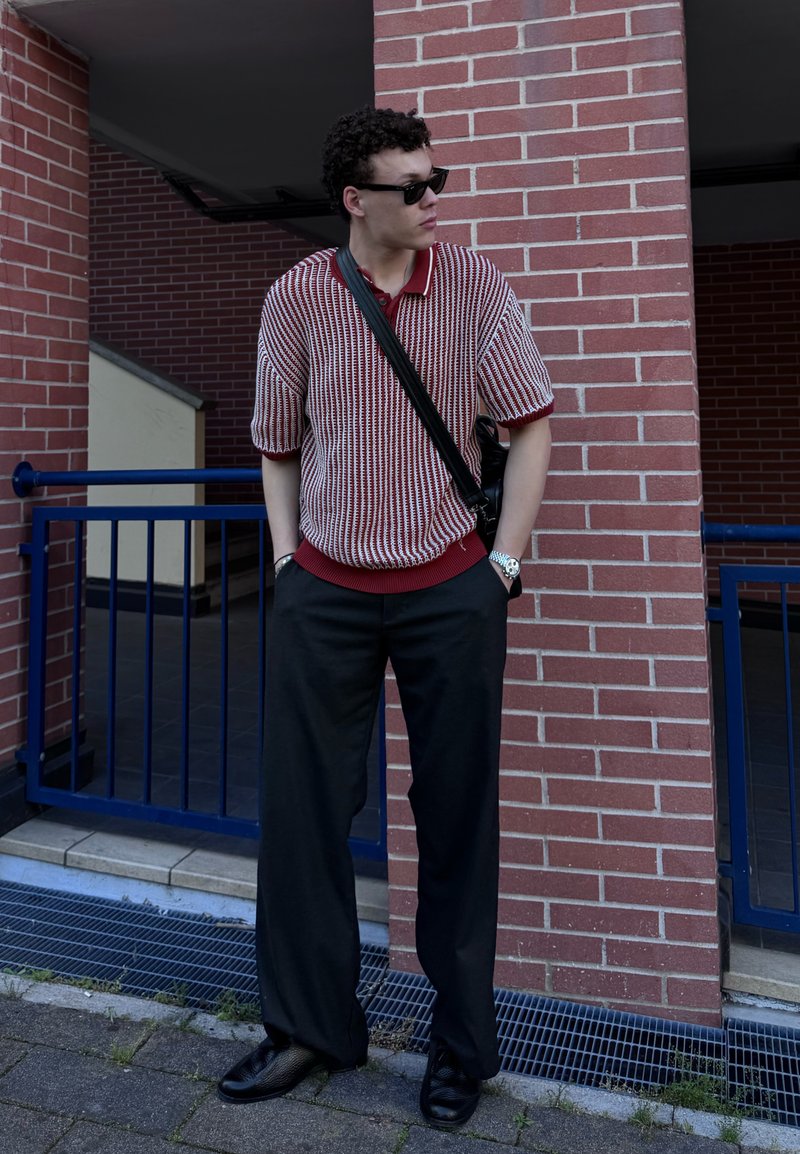 Young man wearing sunglasses, striped short-sleeved shirt, black trousers, and crossbody bag standing against a brick wall near a blue railing.