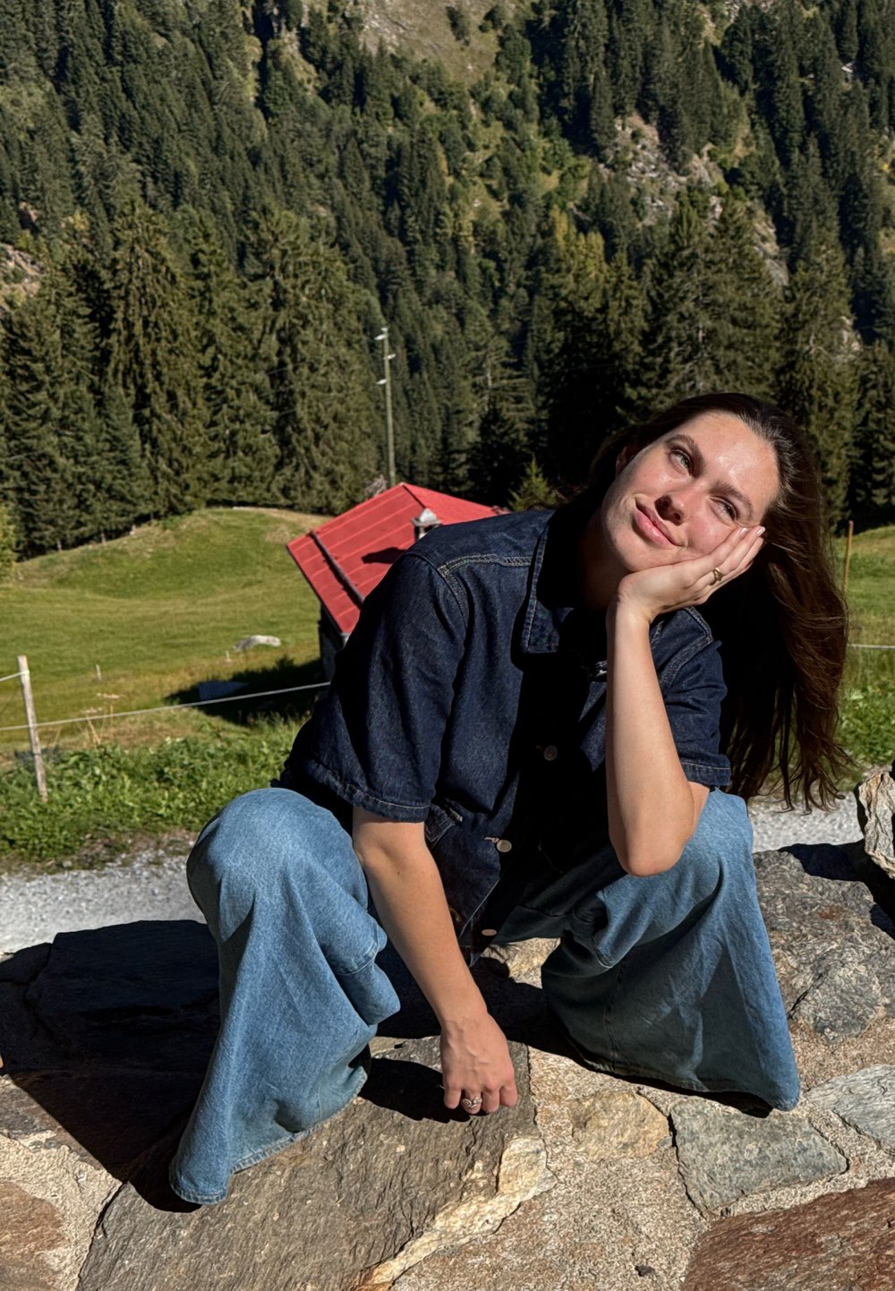 Vrouw in een denimjack en jeans hurkt op een stenen pad, steunt haar hoofd op haar hand, met bosrijke heuvels en een gebouw met een rood dak op de achtergrond.