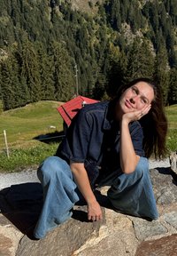 Vrouw in een denimjack en jeans hurkt op een stenen pad, steunt haar hoofd op haar hand, met bosrijke heuvels en een gebouw met een rood dak op de achtergrond.
