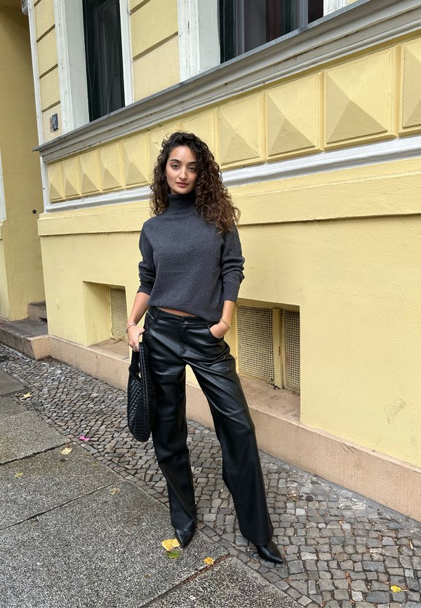 Szürke garbó, magas derekú fekete bőrnadrág és fekete szőtt táska. Macskaköves úton áll, sárga háttérfal előtt.