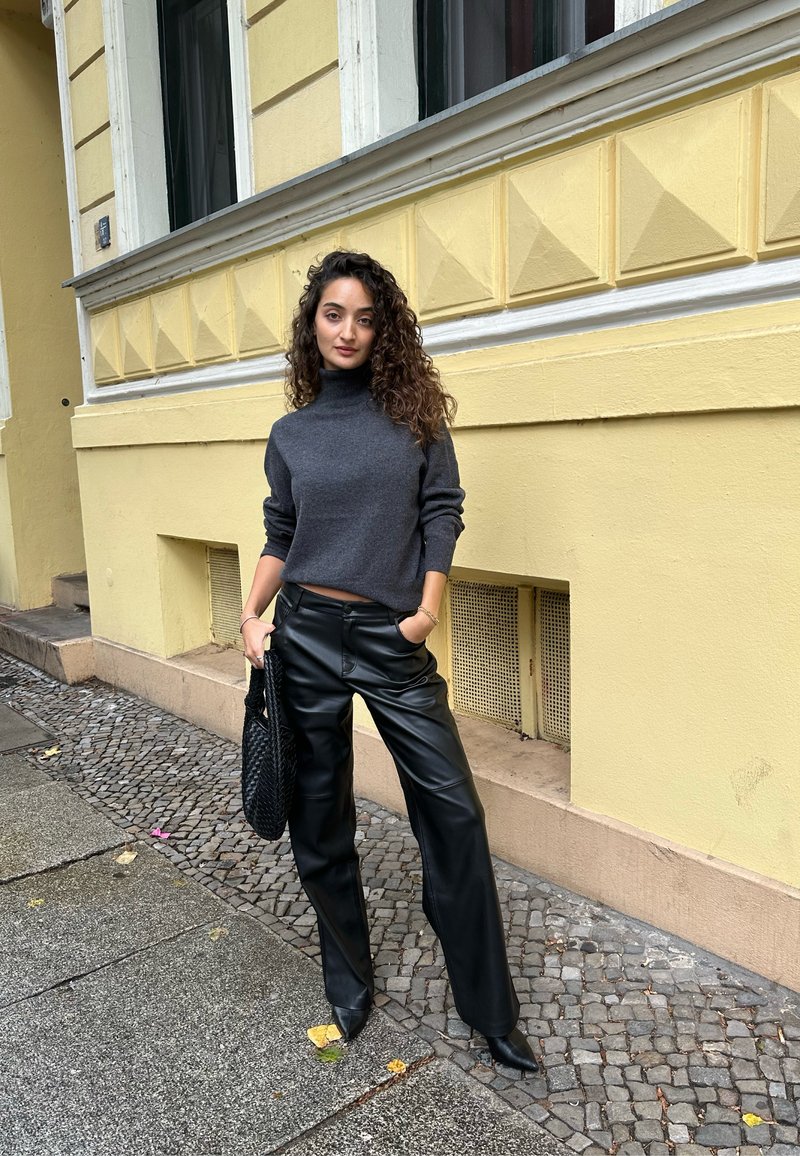 Grauer Rollkragenpullover, hoch sitzende schwarze Lederhose und schwarze geflochtene Handtasche. Stehend auf Kopfsteinpflaster, mit gelber Wand im Hintergrund.
