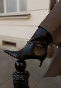 Zwarte leren enkellaars met een puntige neus, slanke hak en een decoratieve riem met een zilveren gesp. Gladde textuur en glanzende afwerking.