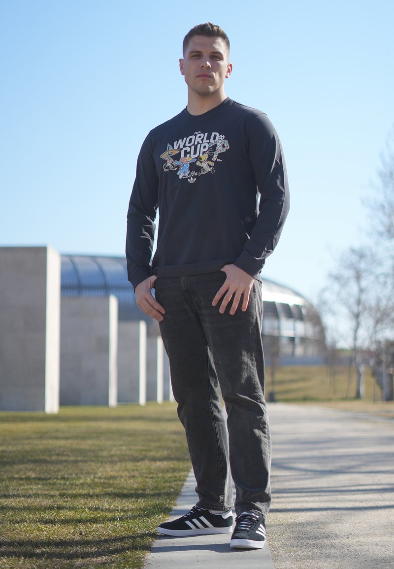 Jeune homme debout en plein air sur un sentier, portant un sweat-shirt sombre de la Coupe du Monde de la FIFA, un jean noir et des baskets Adidas noires par une journée ensoleillée.