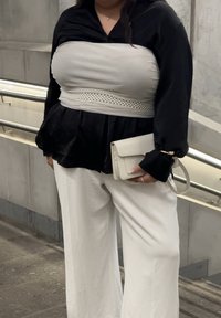 Γυναίκα με μαύρη μπλούζα, μπεζ μπούστo και φαρδιά λευκά παντελόνια, κρατώντας ένα μπεζ τσαντάκι ενώ στέκεται κοντά σε μεταλλική κουπαστή.