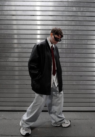 Persona vistiendo una chaqueta negra oversized, camisa blanca, corbata roja, vaqueros claros holgados, zapatillas blancas y gafas de sol oscuras, caminando frente a un fondo de persiana metálica.