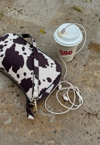 Sac à main imprimé vache, écouteurs blancs branchés sur un gobelet de café à emporter avec un couvercle, posés sur une surface en pierre texturée.