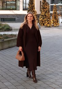 Καφέ ριγέ φόρεμα με φερμουάρ στον λαιμό, μακρύ καφέ παλτό, καφέ μπότες μέχρι το γόνατο και πλεγμένη καφέ τσάντα. Στο φόντο υπάρχουν φωτισμένα αειθαλή δέντρα.