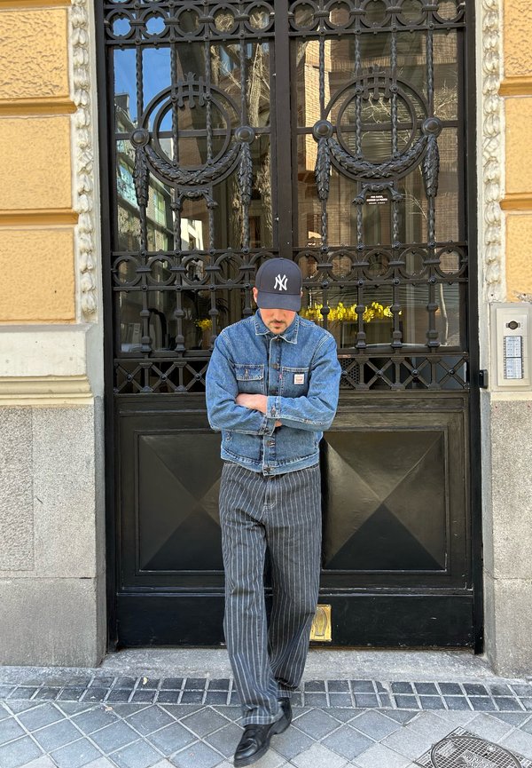 Chaqueta de mezclilla con un tono azul, pantalones grises a rayas y una gorra negra con un logo blanco. De pie frente a una puerta decorativa de metal negro.