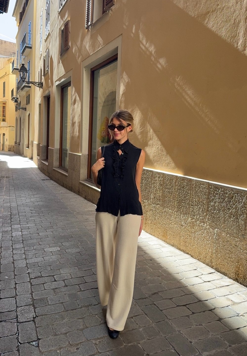 Kobieta w czarnej bezrękawnikowej bluzce i kremowych szerokich spodniach stoi na brukowanej ulicy z żółtymi budynkami i zacienionymi ścianami.