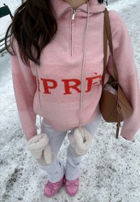 Personne portant un pull rose avec des lettres rouges, un pantalon gris, des chaussures roses, des mitaines blanches suspendues et un sac à bandoulière marron sur un sol enneigé.