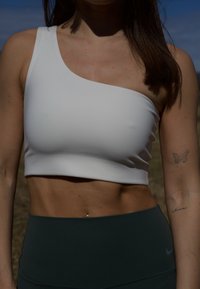 Weißes, einseitiges Cropped-Top aus elastischem Material, kombiniert mit dunkelgrünen, hoch taillierten Leggings, die dezente Markenkennzeichnung am Bein aufweisen.