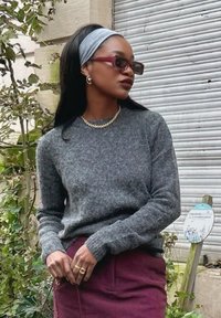 Maglione di lana grigio con scollatura rotonda, abbinato a una gonna in corduroy bordeaux. Gli accessori includono orecchini a cerchio in argento e occhiali da sole rossi.