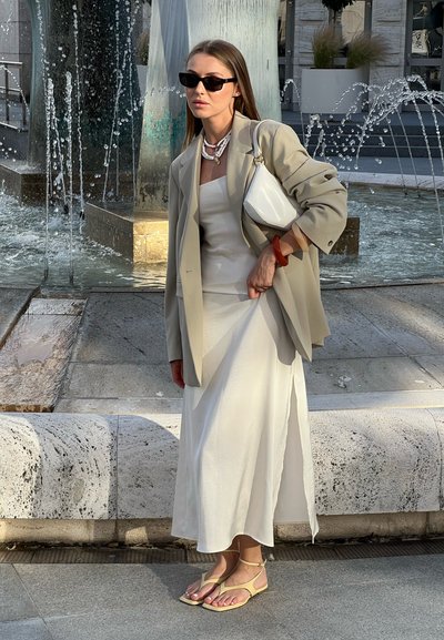 Blazer oversized color beige claro sobre un vestido blanco, combinado con sandalias planas amarillas. Los accesorios incluyen un bolso blanco y collares en capas.
