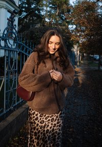 Young woman smiling while buttoning brown fleece jacket, wearing leopard print skirt and burgundy shoulder bag, standing on leafy sidewalk next to fence.
