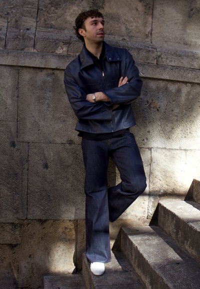 Hombre con cabello rizado, vistiendo una chaqueta de cuero negra y jeans, apoyado contra una pared de piedra junto a unas escaleras al aire libre, mirando de lado.