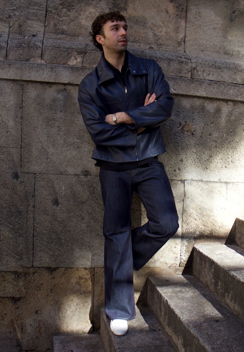 Homme aux cheveux bouclés portant une veste en cuir noire et un jean, appuyé contre un mur en pierre près d'un escalier extérieur, regardant sur le côté.