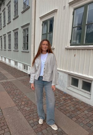 Cárdigan de lino gris claro con un corte relajado, cierre de botones y bolsillos, combinado con jeans acampanados azules desgastados y zapatillas blancas.