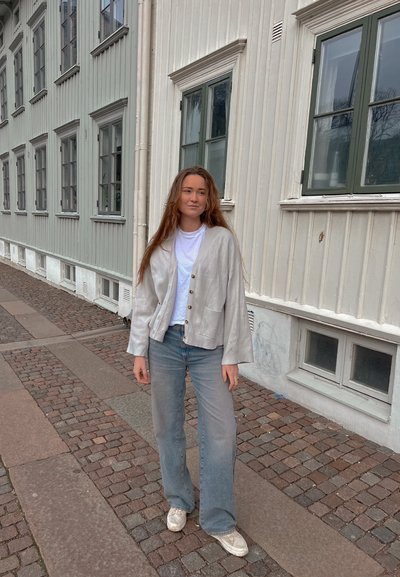 Cárdigan de lino gris claro con un corte relajado, cierre de botones y bolsillos, combinado con jeans acampanados azules desgastados y zapatillas blancas.