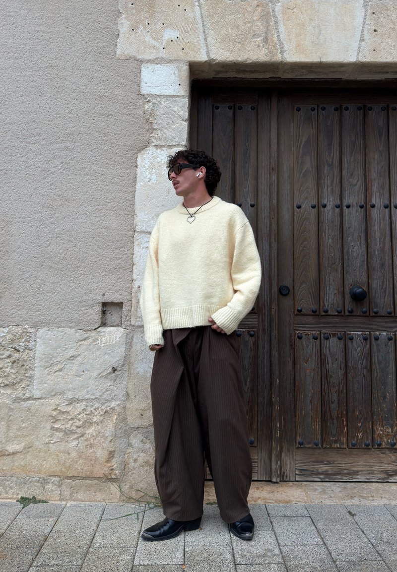 Man wearing cream sweater, loose brown pants, black boots, heart necklace, sunglasses, and earbuds stands against stone wall and wooden door.