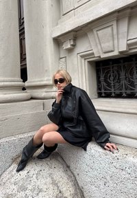 Schwarze, übergroße Lederjacke mit hohem Kragen, kombiniert mit einem schwarzen Minikleid und knielangen schwarzen Stiefeln, sitzend an einer Steinoberfläche.