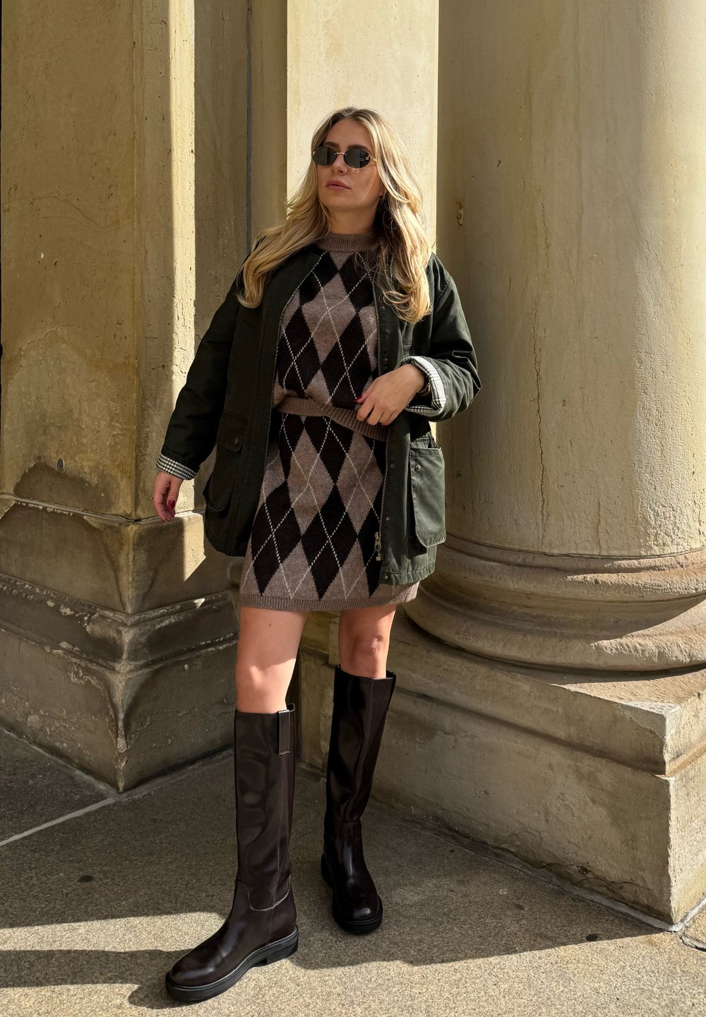 Knielange schwarze Lederstiefel, eine dunkelgrüne Jacke und ein braunes Argyle-Strickkleid mit tailliertem Schnitt. Im Hintergrund sind strukturierte Steinsäulen zu sehen.