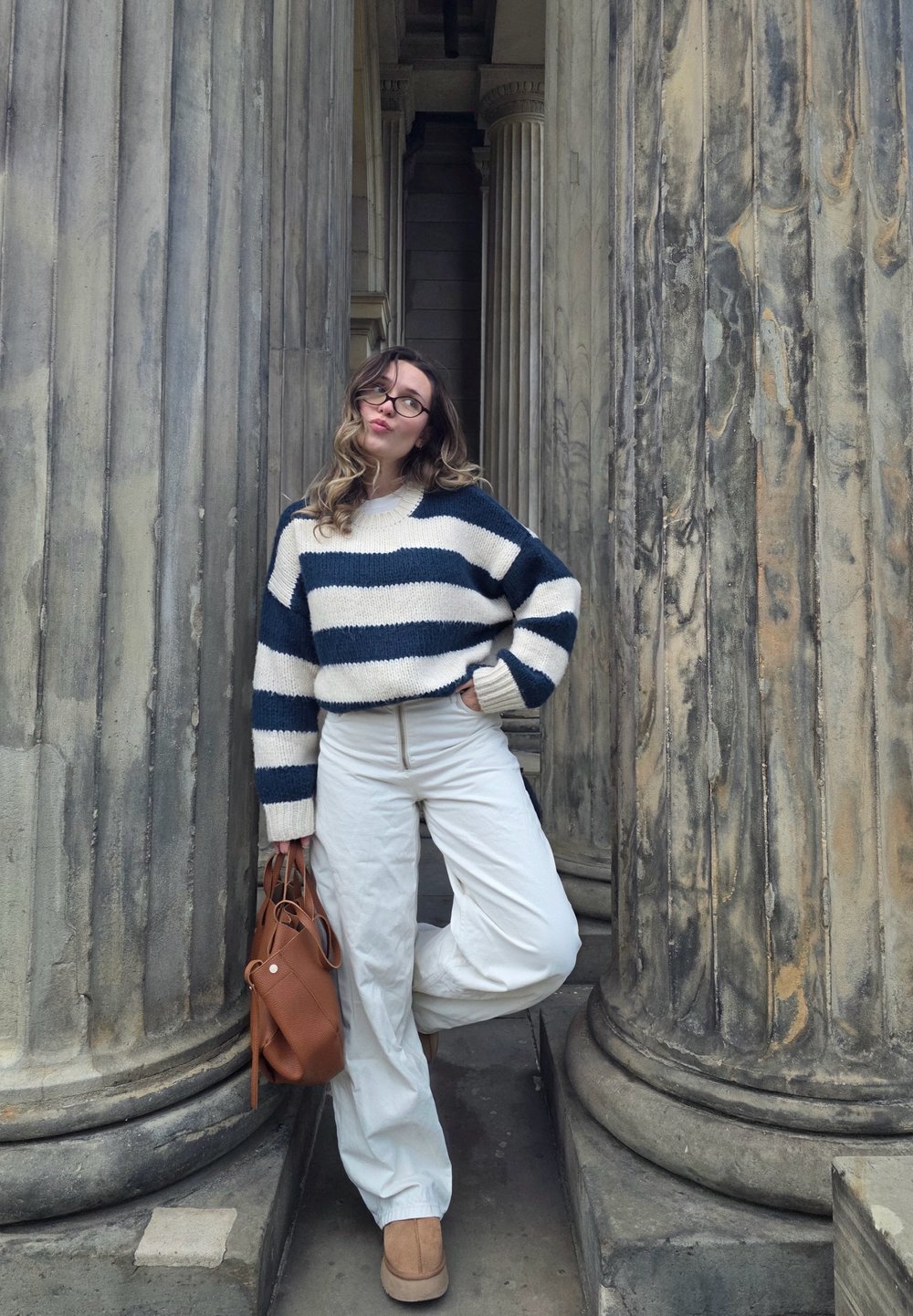 Grobstrickpullover mit marineblauen und cremefarbenen Streifen, weißen Hosen, beigen Stiefeletten und einer braunen Lederhandtasche vor klassischen Säulen.