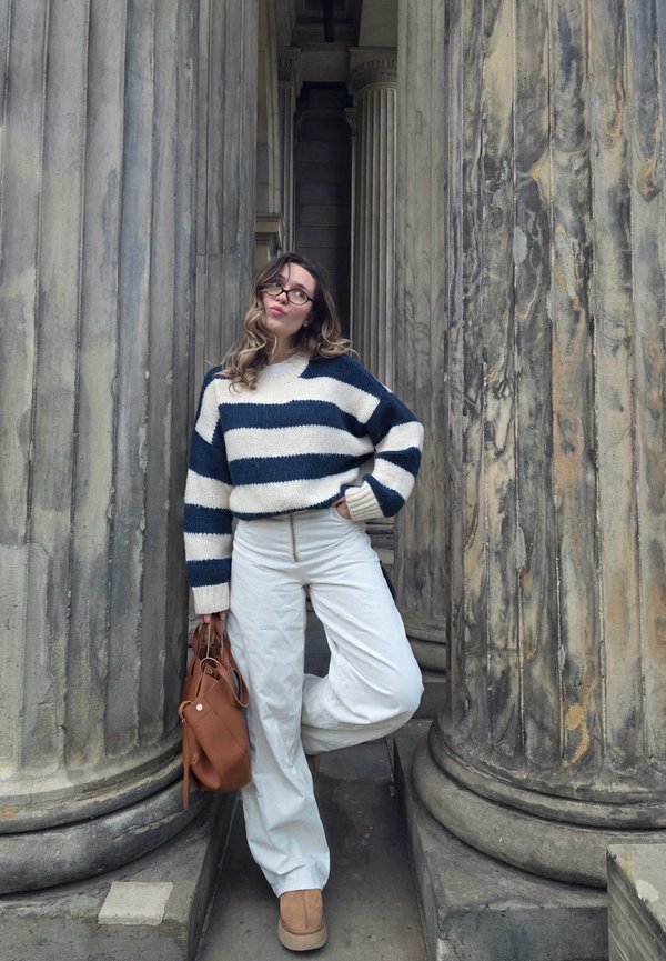 Chunky knit sweater with navy and cream stripes, white trousers, beige ankle boots, and a brown leather bag against classical columns.