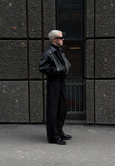 Chaqueta de cuero negra con un diseño estructurado, combinada con pantalones sueltos negros y calzado negro. Cabello corto de color rubio platino y gafas de sol.