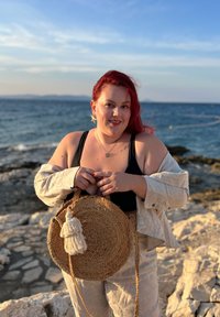 Okrągła, pleciona torba ze słomy z frędzlami, beżowa lniana koszula na czarnym topie, stojąca na skalistym brzegu z morzem w tle.