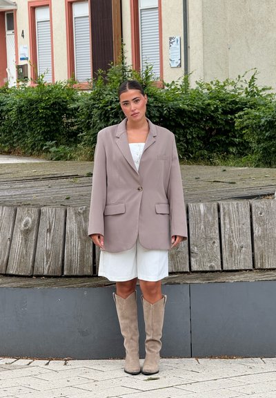 Blazer beige de gran tamaño con solapas muescas y bolsillos frontales, combinado con un vestido blanco y botas de ante beige hasta la rodilla.
