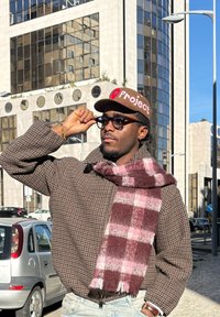 Manteau marron à motif pied-de-poule,  épais foulard écossais bordeaux et rose, casquette de baseball marron avec le texte "Project", lunettes, cadre urbain en plein air.