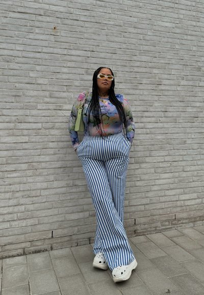 Modelo con una blusa transparente de manga larga con estampado floral, pantalones de pierna ancha a rayas azul y blanca, y zuecos blancos, con un bolso verde.