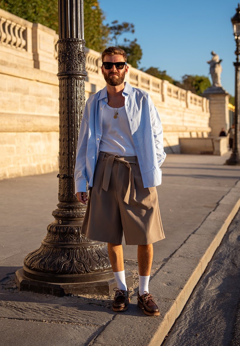 Homme avec barbe et lunettes de soleil debout près d’un lampadaire orné, portant une chemise bleue claire ouverte, un débardeur blanc, un short beige, des chaussettes blanches et des chaussures marron.