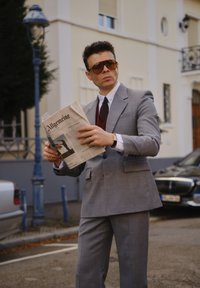 Fato cinza com um acabamento texturizado, combinado com uma camisa de dress branca e uma gravata com padrão. Segurando um jornal intitulado "Allgemeine." Usando óculos de sol oversized.