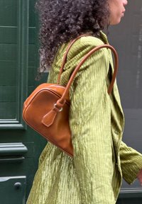 Bruine leren handtas met een afgeronde vorm, ritssluiting en een lange schouderband, vastgehouden door een persoon in een gemêleerde olijfgroene jas.