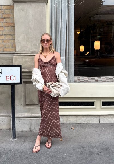 Vestido marrón de lunares con un corsé ajustado y tirantes finos, combinado con una chaqueta blanca de gran tamaño, sosteniendo un bolso beige.