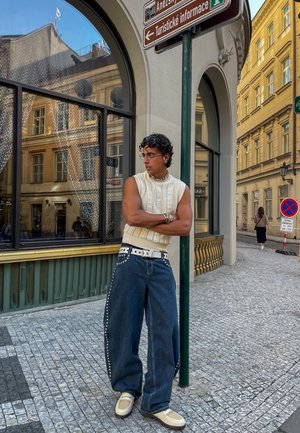 Top sin mangas de punto blanco, jeans holgados de mezclilla oscura con tachuelas, cinturón blanco, zapatillas beige, gafas y un collar plateado. Entorno urbano.